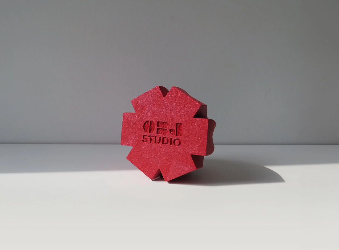 Red geometric object with OBJ STUDIO logo, 3D printed from plant-based PLA and styled in natural light.